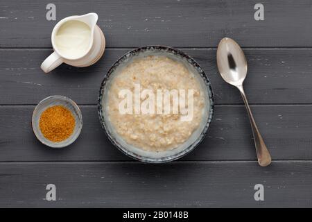 Eine nahrhafte Porridschüssel, eine kleine Schüssel mit braunem Zucker, eine Kanne Milch und einen Löffel, auf dunklem Holzhintergrund Stockfoto