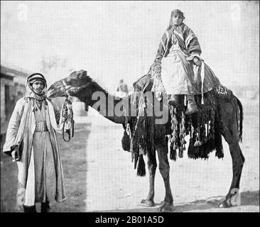 Syrien: Beduinenfrau auf einem Kamel geführt von ihrem Mann, c. 1905. Die Beduinen (aus dem arabischen badawi, pl. badawiyyūn) gehören zu der überwiegend wüstenbewohnten arabischen Volksgruppe. Konkret bezieht sich der Begriff nur auf die "Kamelzüchtiger" Stämme, aber aufgrund wirtschaftlicher Veränderungen sind viele mittlerweile angesiedelt oder züchten Schafe. Auch aufgrund sprachlicher und kultureller Veränderungen wird der Begriff heute häufig auf vielfältige Weise entweder für Araber im Allgemeinen oder für Wüstenbewohner oder Nomaden verwendet. In Syrien leben die Beduinen überwiegend in der Wüste östlich und südlich des Landes. Stockfoto