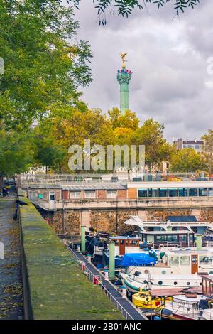 Paris, Frankreich - 11. November 2019: Der obere Teil des Bastille-Denkmals hinter Bäumen, vom Bourdon Boulevard. Boote im Hafen von Arsenal Stockfoto