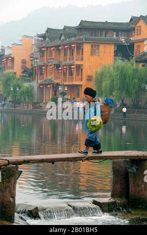 China: Tujia-Frau auf der alten Holzbrücke über den Tuo-Fluss, Fenghuang, Provinz Hunan. Fenghuang ist Chinesisch für Phoenix und bezieht sich auf den mythischen heiligen feuervogel, der in den Mythologien der Peter, Griechen, Römer, Ägypter, Chinesen, Und (nach Sanchuniathon) die Phönizier. Die Legende besagt, dass zwei Phönixe, die die Stadt entdeckten, einige Zeit über dem Kopf schwebten, bevor sie widerwillig wegflogen. Die Stadt Fenghuang ist eine gut erhaltene antike Stadt, die angeblich aus dem Jahr 248 v. Chr. stammt. Hier leben die Minderheiten Miao und Tujia. Stockfoto