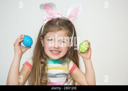 Süßes kleines Mädchen, das bunte Ohren trägt und gemalte Ostereier hält und auf weißem Hintergrund lächelt. Ostertag. Glückliches Baby, das Urlaub genießt. Stockfoto