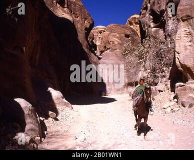 Jordanien: Junge Reiter im Siq (Schacht), der zur antiken Stadt Petra führt. Der Siq oder al-Siq ist der Haupteingang zur antiken Stadt Petra im südlichen Jordanien. Die dunkle, schmale Schlucht (an einigen Stellen nicht mehr als 3 Meter breit) windet sich etwa 1,5 km und endet bei Petras aufwändigster Ruine, Al Khazneh (die Schatzkammer). Petra wurde im 4. Jahrhundert v. Chr. von den nabatäischen Arabern als Stadt gegründet und verdankt seine Geburt und seinen Wohlstand der Tatsache, dass es der einzige Ort mit klarem und reichlich Wasser zwischen den Hidschas-Handelszentren Mekka und Medina und Palästina war. Stockfoto