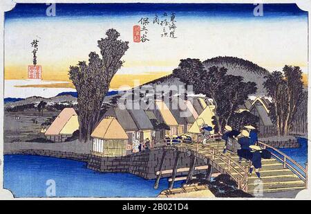 Japan: Station 4: Hodogaya Station und Shinkame Bridge. Ukiyo-e-Holzschnitt aus der Serie „Three Stations of the Tôkaidô Road“ von Utagawa Hiroshige (1797-1858), um 1834. Hodogaya: Eine Brücke über einen Bach, über den zwei Kolien ein geschlossenes Kago in Richtung eines Dorfes auf der gegenüberliegenden Uferseite tragen; hinter dem Dorf erhebt sich ein niedriger hölzerner Hügel. An dieser Landstation hatten die Reisenden eine Entfernung von 34 km von Edo zurückgelegt und waren gut in Reisestimmung. An der Brücke steht ein Restaurant, das Soba (Buchweizennudeln) serviert, mit Kellnerinnen, die Reisende einladen. Stockfoto