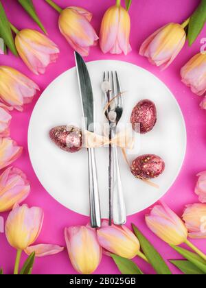 Schöne luxuriöse easter Tischbestecke wie Messer, Gabel mit bunten Tulpen und goldbemalten Eiern mit Glitzern auf rosa Oberfläche. Weihnachtshintergrund, Grußkarte, Nahaufnahme Stockfoto