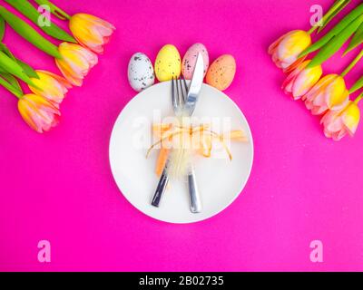 Festliches ostertischbesteck mit Messer, Gabel, verziert mit Feder, Schleife, mehrfarbigen Eiern, gelben Tulpenblumen auf pinkfarbenem Federferienhintergrund. Draufsicht Stockfoto