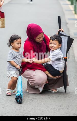 Vertikale Ansicht einer geschäftigen muslimischen Mutter in roten Hijab Squat nach unten zu halten und zu unterstützen ihr weinendes Kind auf der Straße. Singapur. Stockfoto