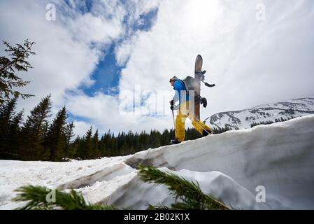 Blick auf die Snowboarder in hellem Skianzug mit Snowboard auf dem Rücken und aufsteigender Schneedecke bis zum Berggipfel. Abenteuer in den Bergen. Wolken am blauen Himmel auf dem Hintergrund. Ansicht mit niedrigem Winkel Stockfoto
