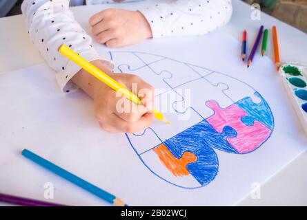 Ein Kind zeichnet buntes Herz mit Puzzle auf weißem Hintergrund als Symbol für Autismus, mit bunten Bleistiften und Aquarellen. Stockfoto