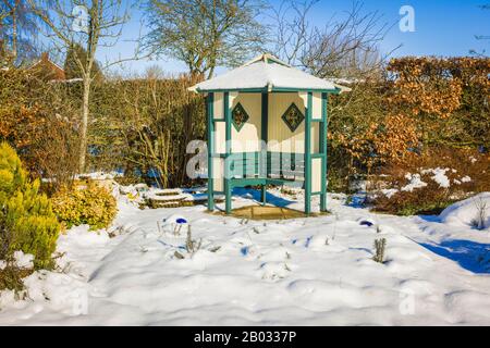 Winterschneefall verwandelt den Vergnügungsgarten, verbirgt Unkraut und verleiht der saisonalen Grauigkeit Licht. Ein Arbour ist an sonnigen Tagen immer noch erreichbar und nützlich Stockfoto