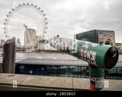 Ein mit Aufklebern überdecktes Touristenteleskop, das in der Hintergrundunschärfe über die Themse in Richtung London Eye blickt. Stockfoto