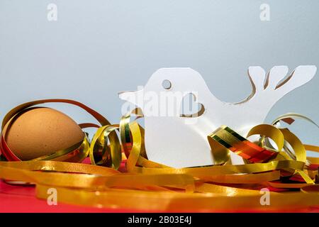 Papiergeschnittener weißer Vogel mit Herz und braunem Ei zwischen glänzenden gelben Bändern auf roter Pappoberfläche auf grauem Grund. Mit Kopierbereich Stockfoto