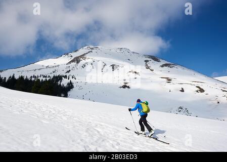 Männlicher Wanderer, Tourist in farbenfroher Kleidung mit Rucksackwandern auf Skiern auf einem schneebedeckten Hügel vor hellblauem Himmel und schöner Bergwelt an frostigen sonnigen Tagen. Winter-aktives Lifestyle-Konzept. Stockfoto