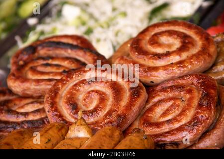 Gegrillte Spiralwürste auf einem Teller mit Grün und Gemüse im Hintergrund Straßennahrung Stockfoto