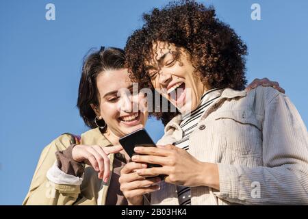 Interracial Paar von zwei jungen schönen Frauen, die beim betrachten des Telefons, des Konzepts der Technologie und der rassischen Vielfalt lachen Stockfoto