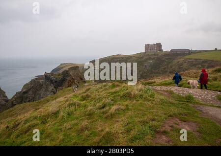 Zwei Hundegänger, die das nasse Wetter am Glebe Cliff in Richtung Tintagel Castle Ruins mit Camelot Castle Hotel auf dem Clifftop in Cornwall, Großbritannien, braten. Stockfoto