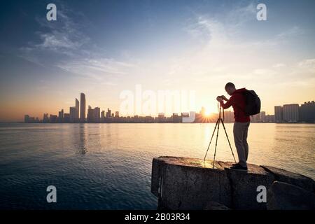 Fotograf mit Kamera auf Stativ fotografiert bei Sonnenaufgang die Skyline der Stadt. Abu Dhabi, Vereinigte Arabische Emirate. Stockfoto