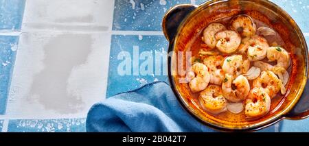 Fisch und Meeresfrüchte Vorspeise aus frischen Scampi und Knoblauch, gewürzt mit Kräutern und Gewürzen, die in einem Panorama-Banner auf einer blau-weiß gefliesten Oberfläche zur Seite serviert werden Stockfoto