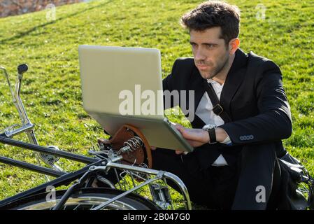 Mann im Anzug und Fahrrad schaut auf seinen Computer, der auf dem Gras sitzt Stockfoto