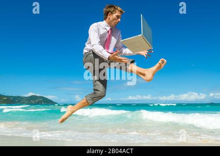 Energiegeladener Büroangestellter springt mit seinem Laptop an einem leeren tropischen Strand entlang Stockfoto