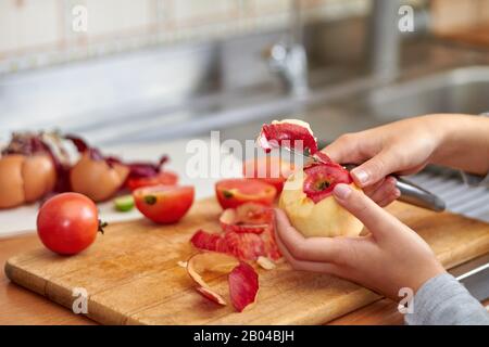 Das Mädchen schneidet eine Schale mit einem Messer von einem roten apfel. Lebensmittelabfälle und -Reste bei der Zubereitung von Speisen in der Küche. Nahaufnahme, selektiver Fokus Stockfoto