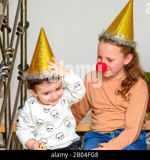 Porträt des Teenagermädchens mit einer Clownse und einem Hut, der sich mit dem kleinen Bruder im Partyhut auf der Purim- oder Geburtstagsfeier lustig macht. Stockfoto