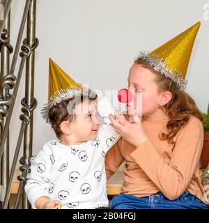 Porträt des Teenagermädchens mit einer Clownse und einem Hut, der sich mit dem kleinen Bruder im Partyhut auf der Purim- oder Geburtstagsfeier lustig macht. Stockfoto