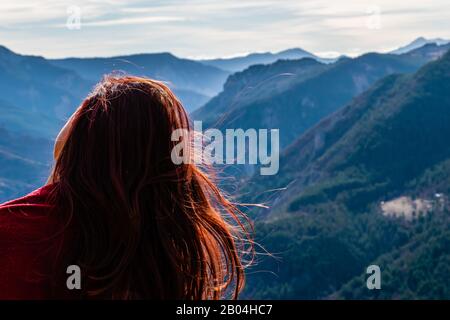 Nahansicht einer jungen Rotkopffrau mit gekipptem Kopf und langem Haar flatternd vor der Bergtallandschaft von hinten mit Stockfoto