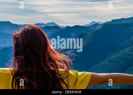 Nahansicht einer jungen Rotkopffrau vor den hochgezogenen Berglandschaftsarmen und von hinten flatternde Haare ohne Gesicht auf sonnigem w Stockfoto