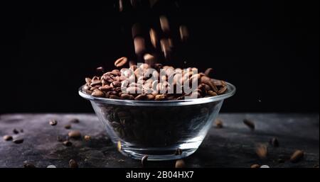 Gebratene Kaffeebohnen, die in einer Glasschale auf schwarzem Grund fallen. Stockfoto