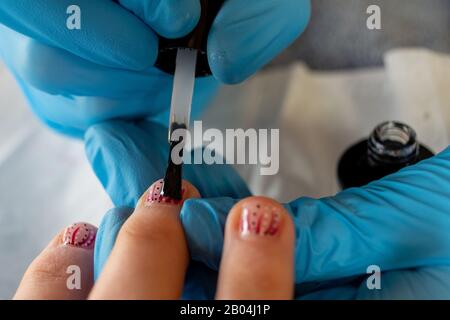 Nahaufnahme von klarem polnisch, das auf kurz dekorierte Nägel aufgetragen wird. Manikurist trägt blaue Handschuhe. Stockfoto