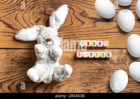 Gefülltes Häschen mit der Botschaft "Hapy Easter" auf Deutsch und vielen Eiern auf Holzhintergrund Stockfoto