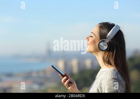 Seitenansicht Portrait eines zufriedenen Mädchens, das Musik hört, Kopfhörer trägt und das Handy im Freien in der Stadt hält Stockfoto