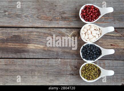 Verschiedene Sorten Bohnen: Baby lima, schwarz klein, Adzuki und Mang in weißen Keramiklöffeln (Vorspeisen) auf Holzgrund, selektives lwl Stockfoto