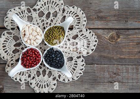 Verschiedene Sorten Bohnen: Baby lima, schwarz klein, Adzuki und Mang in weißen Keramiklöffeln (Vorspeisen) auf Holzgrund, selektives lwl Stockfoto