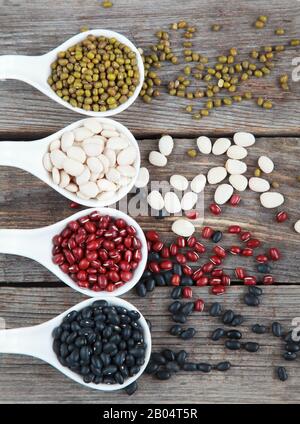 Verschiedene Sorten Bohnen: Baby lima, schwarz klein, Adzuki und Mang in weißen Keramiklöffeln (Vorspeisen) auf Holzgrund, selektives lwl Stockfoto