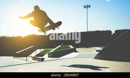 Silhouette des Skaterspringens auf der Rampe im Stadtpark - Junger Mann, der Tricks und Fähigkeiten mit Skateboard bei Sonnenuntergang ausführt - Jugend, Extremsportkonzept - Stockfoto