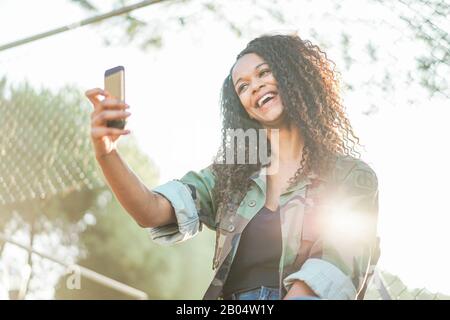 Glückliche afrikanerin spielt mit Smartphone-Kamera im Stadtpark im Freien - Afro-Mädchen hat Spaß, Medien im sozialen Netzwerk mit Handy zu teilen - Neu Stockfoto