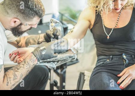 Professioneller bärtiger Tattoo-Künstler, der in seinem eigenen Tintenstudio an der jungen blonden Frau - Tattoer bei der Arbeit - Das Kunst- und Hauttrend-Konzept - Focu tätowiert Stockfoto