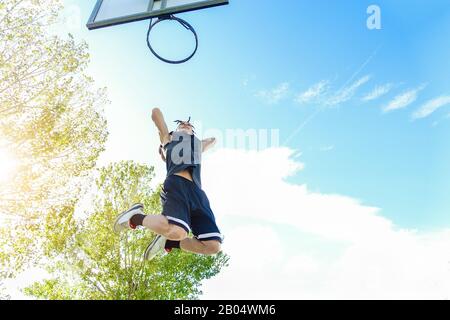 Junge Dreadlocks Spieler, die im städtischen Basketballfeld mit Rückensonnenlicht trunkeln - Starker Athlet, der Slam-Dunk an der Universität ausführt - Sport und sc Stockfoto