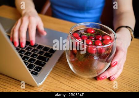 Ein Mädchen in einem blauen Kleid sitzt in einem Café, entspannt Filme, Fernsehsendungen auf einem Laptop und trinkt köstlichen Tee mit roten Beeren. Nahaufnahme. Stockfoto