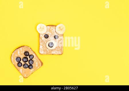 Lustige Erdnussbuttertoasts mit Herzform und lustigen Gesicht für Kinder Frühstück auf gelbem Hintergrund. Draufsicht mit Kopierbereich Stockfoto