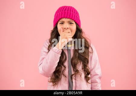 Kleine Mädchen fangen Husten. In diesem Winter keine Grippe. Tragen Sie warme Kleidung in der kalten Jahreszeit. Winterferienaktivität. Kleiner pinkfarbener Hintergrund. Modischer Stil für Kinder. Strickmütze und Puffer-Mantel. Wintergrippe. Stockfoto