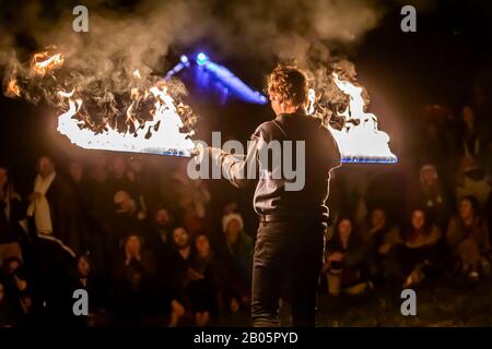 Ein Mann wird während einer Feuerwehr-Tanzroutine nachts brennende Schwerter halten sehen, wobei verschwommene Menschen im Hintergrund beim multikulturellen Erdfest zusehen Stockfoto