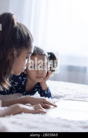 Ein kleines Mädchen mit schwarzen Augen sieht ihre Schwester an, die einen Finger auf ein Buch zeigt. Ein dunkelhäutiges Baby aus dem Nahen Osten sieht ihre Schwester an Stockfoto