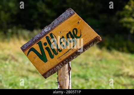 Ein Nahselektiver Fokusschuss eines verzerrten handgefertigten Holzschildes mit der Aufschrift "Village", das an einem Holzpfosten auf einem Zeltplatz mit unscharfem grünen Hintergrund befestigt ist Stockfoto