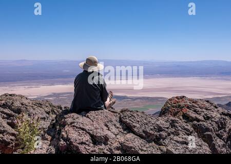 Eine junge Frau in einem Strohhut, die auf einer Klippe mit Blick auf die Alvord Desert sitzt. Steens Mountains im Süden von Oregon Stockfoto