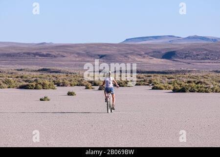 Ein Mädchen im Teenager-Alter, das auf einem playa von Alvord vor Steens Bergen im Hintergrund mit dem Fahrrad unterwegs ist Stockfoto