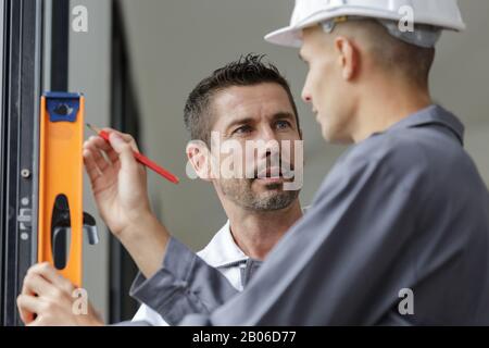 Zwei Erbauer sprechen, während sie die Wasserwaage gegen Fenster halten Stockfoto