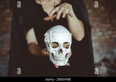 Eine Hexe hält einen menschlichen Schädel in einem schwarzen Mantel in einem alten Gebäude Stockfoto