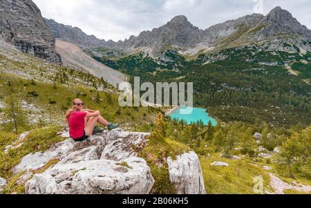 Junge Frau, Wanderer auf Felsen sitzend und mit Blick auf den türkisgrünen Sorapisser See und die Berglandschaft, die Dolmen, Belluno, Italien Stockfoto